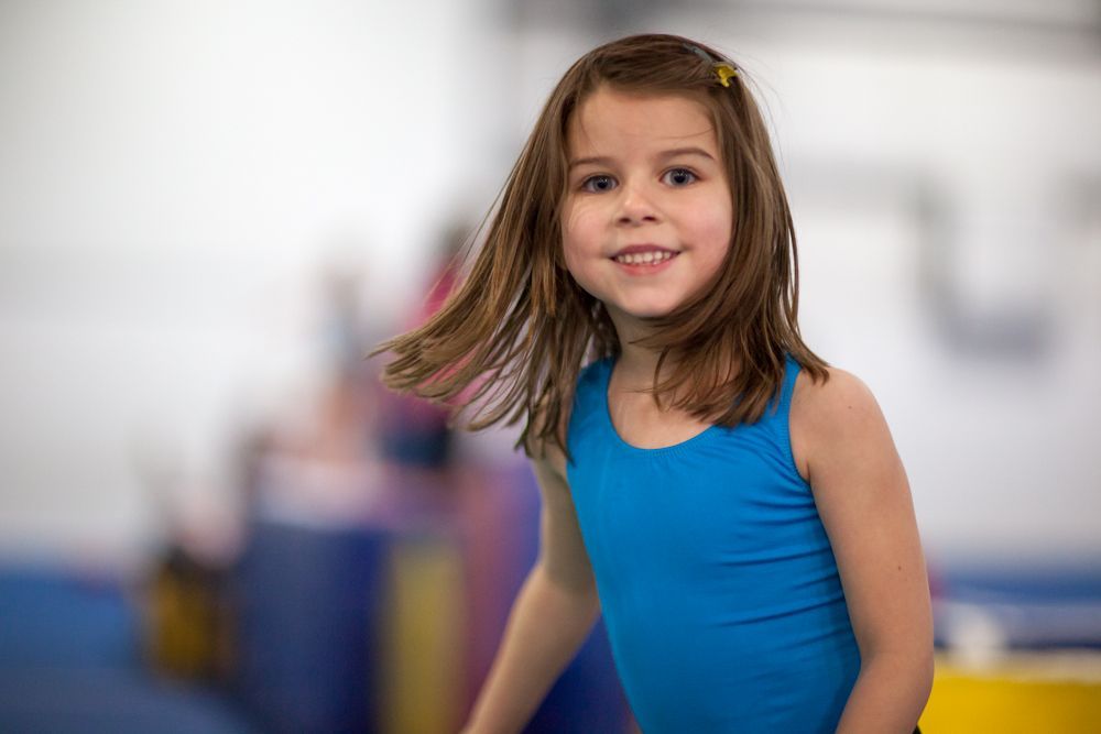 A little girl in a blue tank top is standing on a balance beam in a gym.