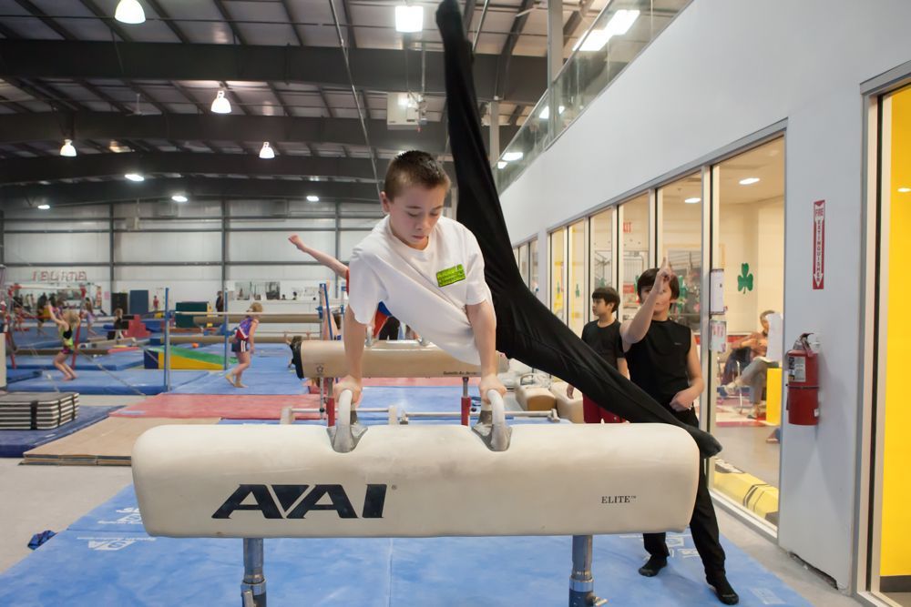 A young boy is doing a split on a pommel horse in a gym.