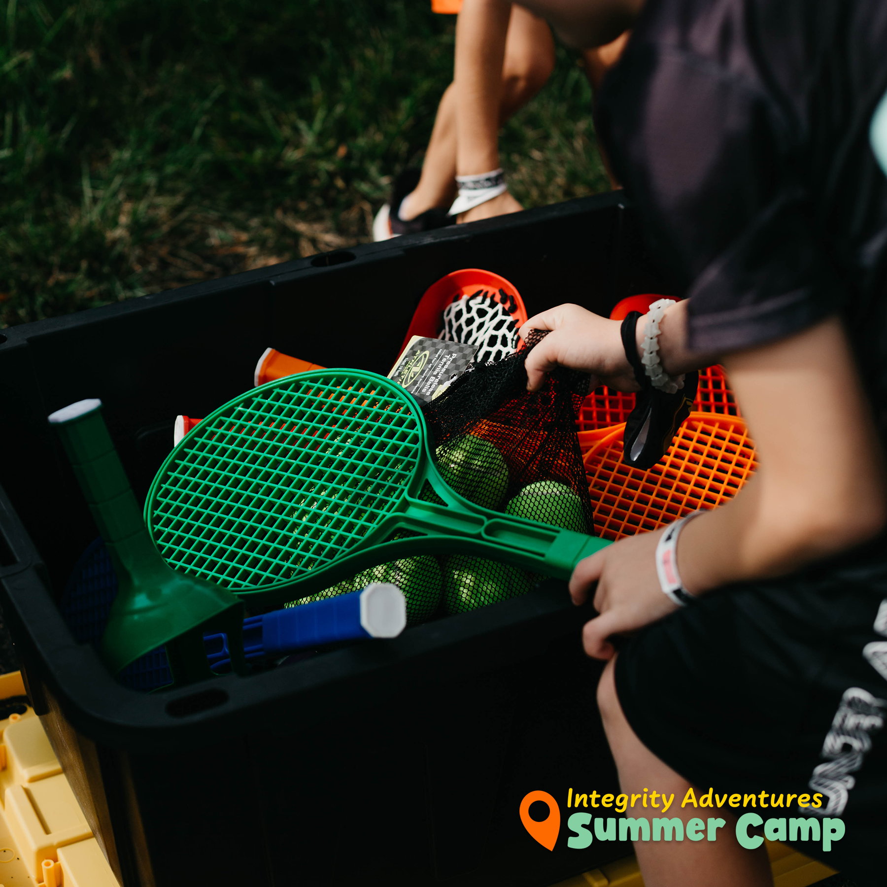 A boy is playing with a green tennis racquet in a box that says integrity adventures summer camp