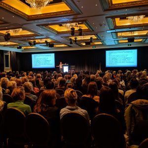 A large group of people are sitting in a large room watching a presentation.
