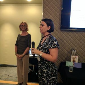 Two women are standing in front of a screen and one is holding a microphone