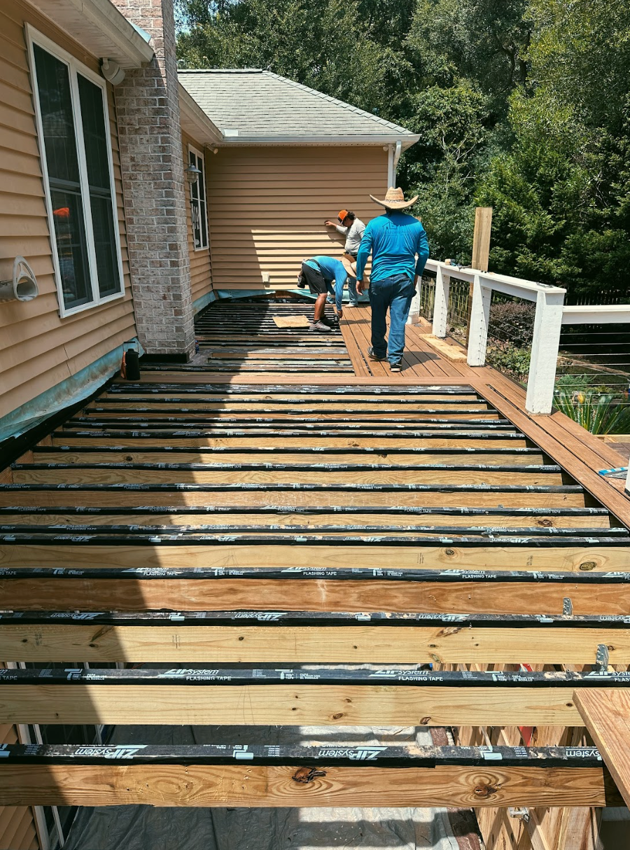 Construction workers building a deck.  Yellow house, wood planks, blue shirts, sunny day, and railing.