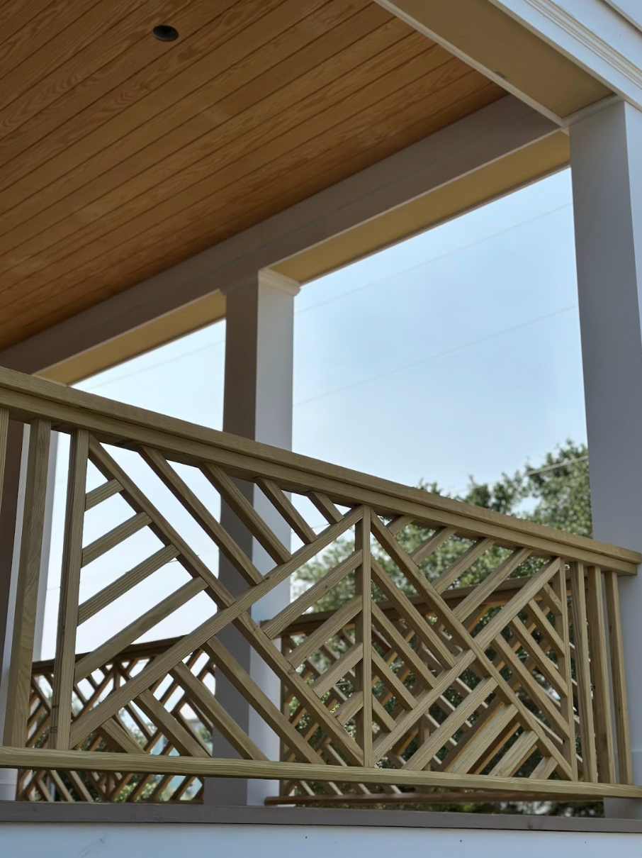 A beige, geometric wooden railing on a porch with white columns, looking out at the sky.