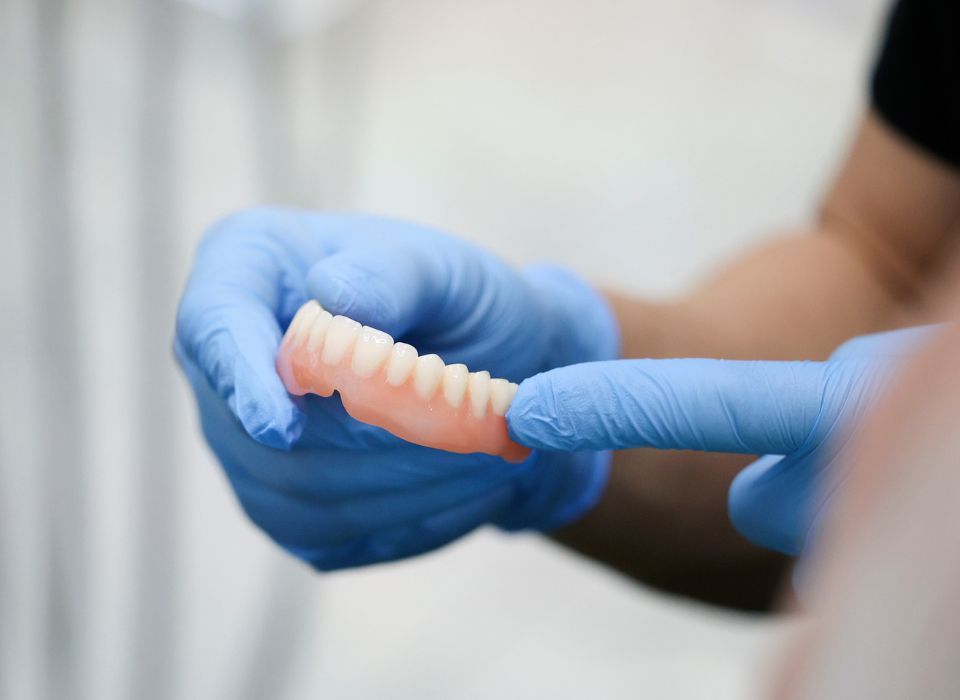 A person wearing blue gloves holds a set of dentures, showing the pink gum and white teeth.