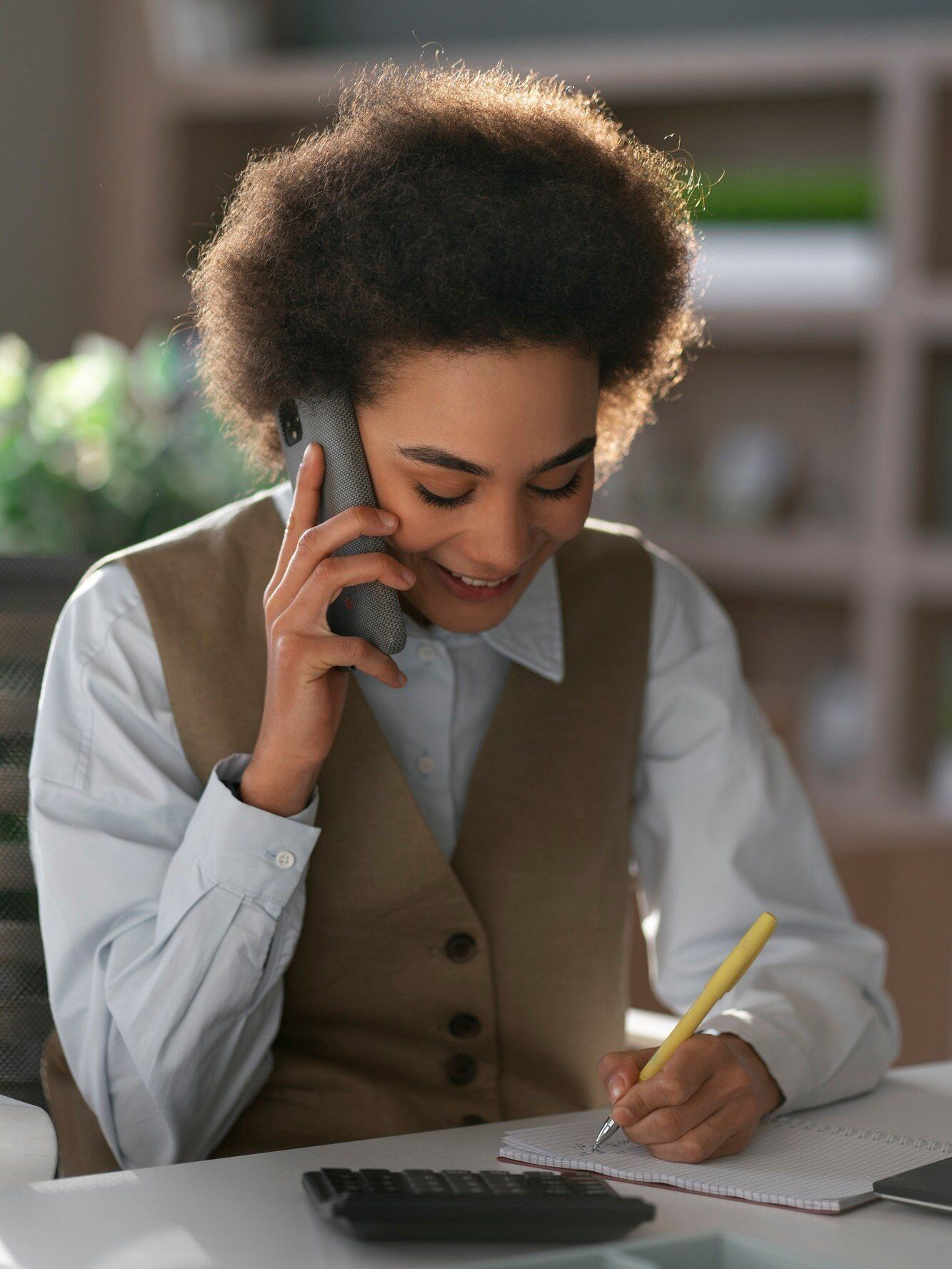 A woman is sitting at a desk talking on a cell phone and writing in a notebook.