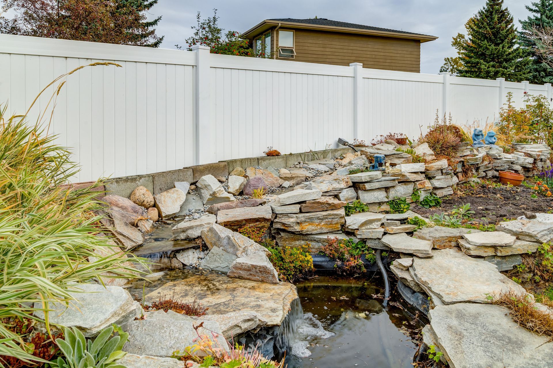 Backyard with a rock waterfall and pond, enclosed by a white fence.