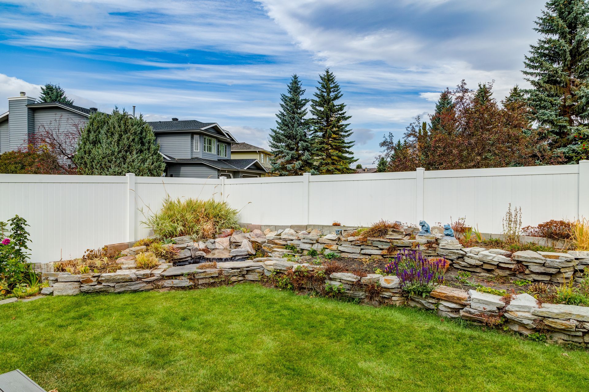 Backyard garden with layered stone beds, white fence, green lawn, and autumn trees under a blue sky
