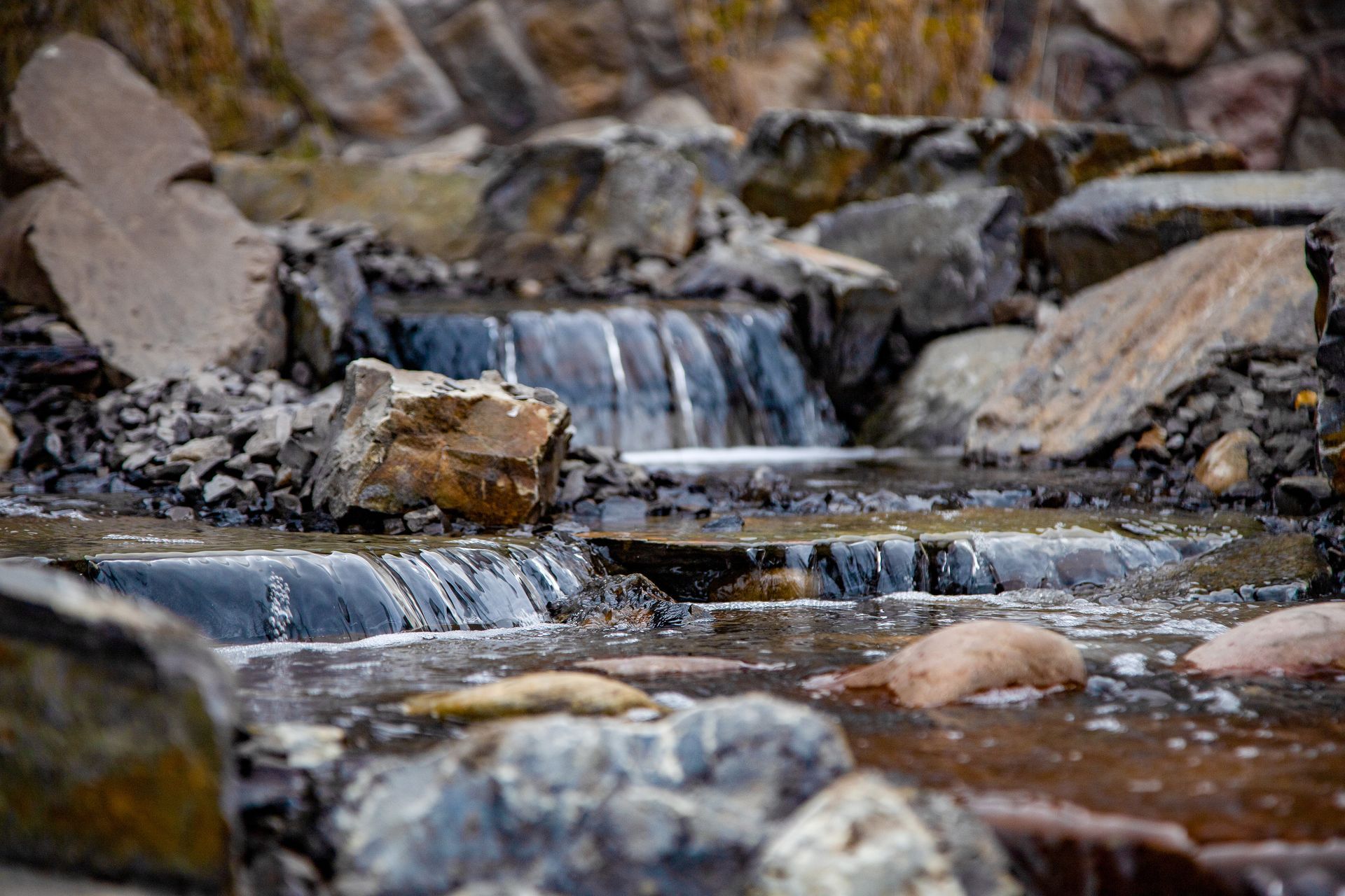 Small waterfall flowing over rocks in a shallow stream, with mossy stones and blurred background