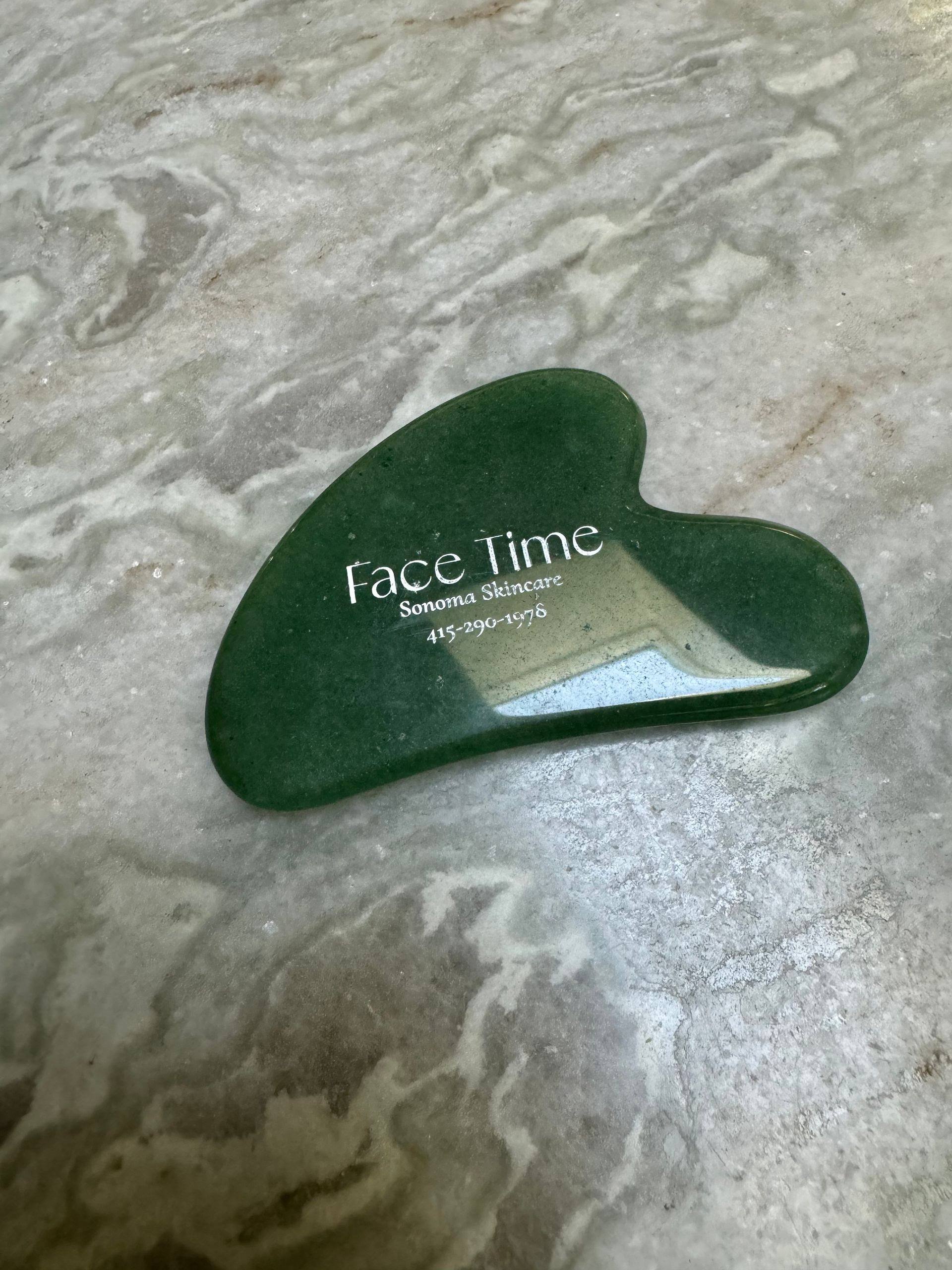 A green heart shaped gua sha is sitting on a marble surface.