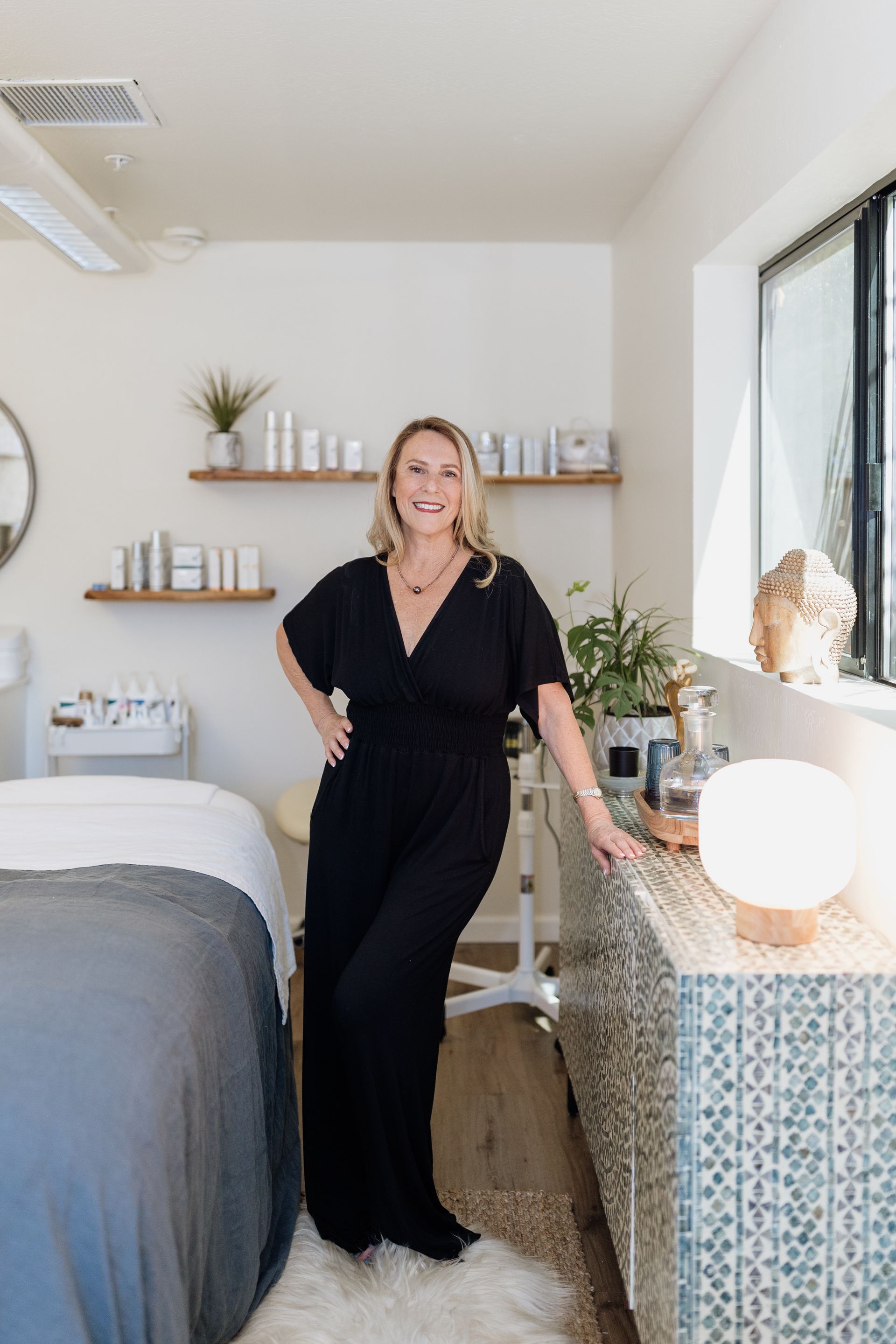 A woman in a black jumpsuit is standing in a room next to a bed.
