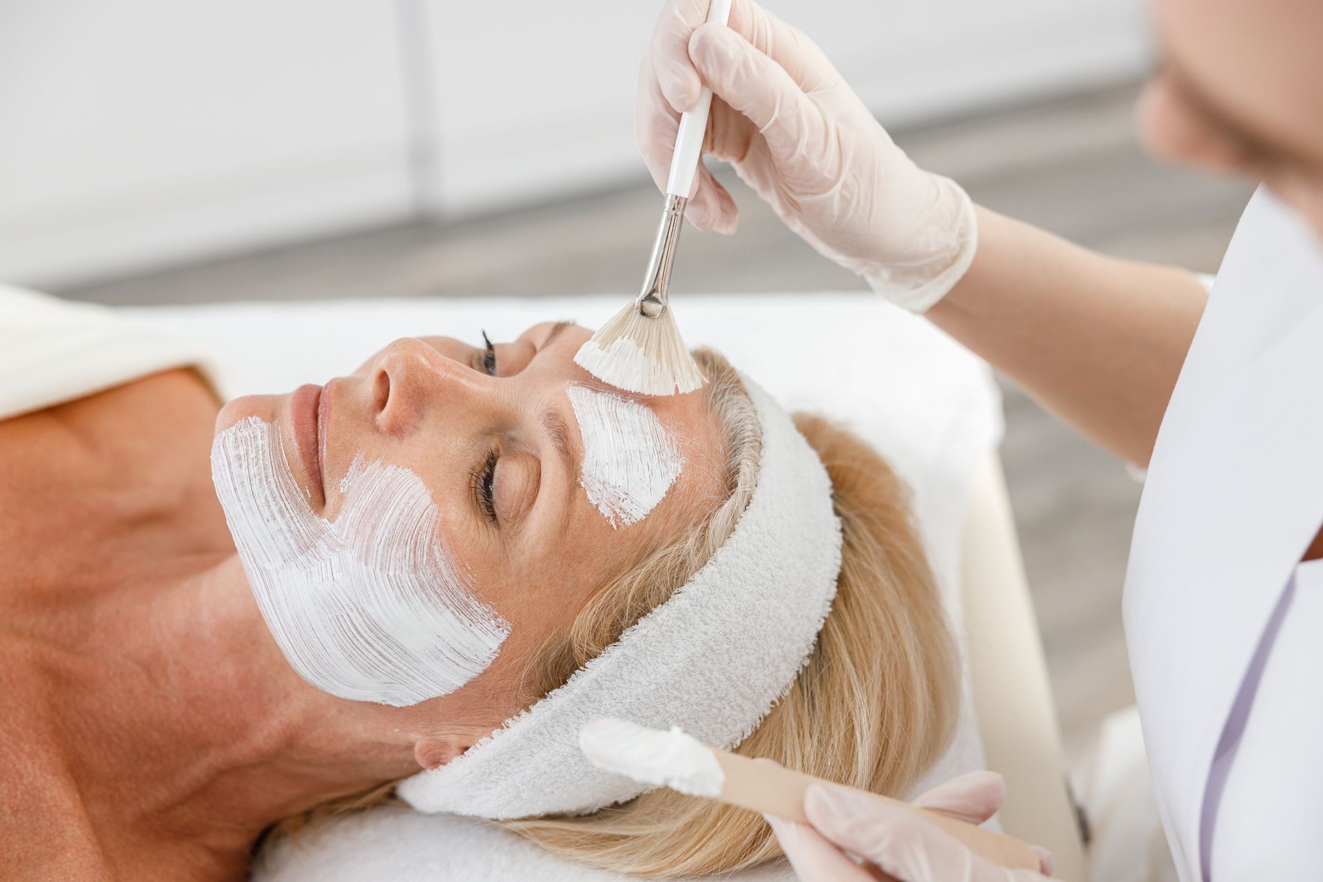 A person wearing gloves applies a white facial mask to a client's face using a small brush.