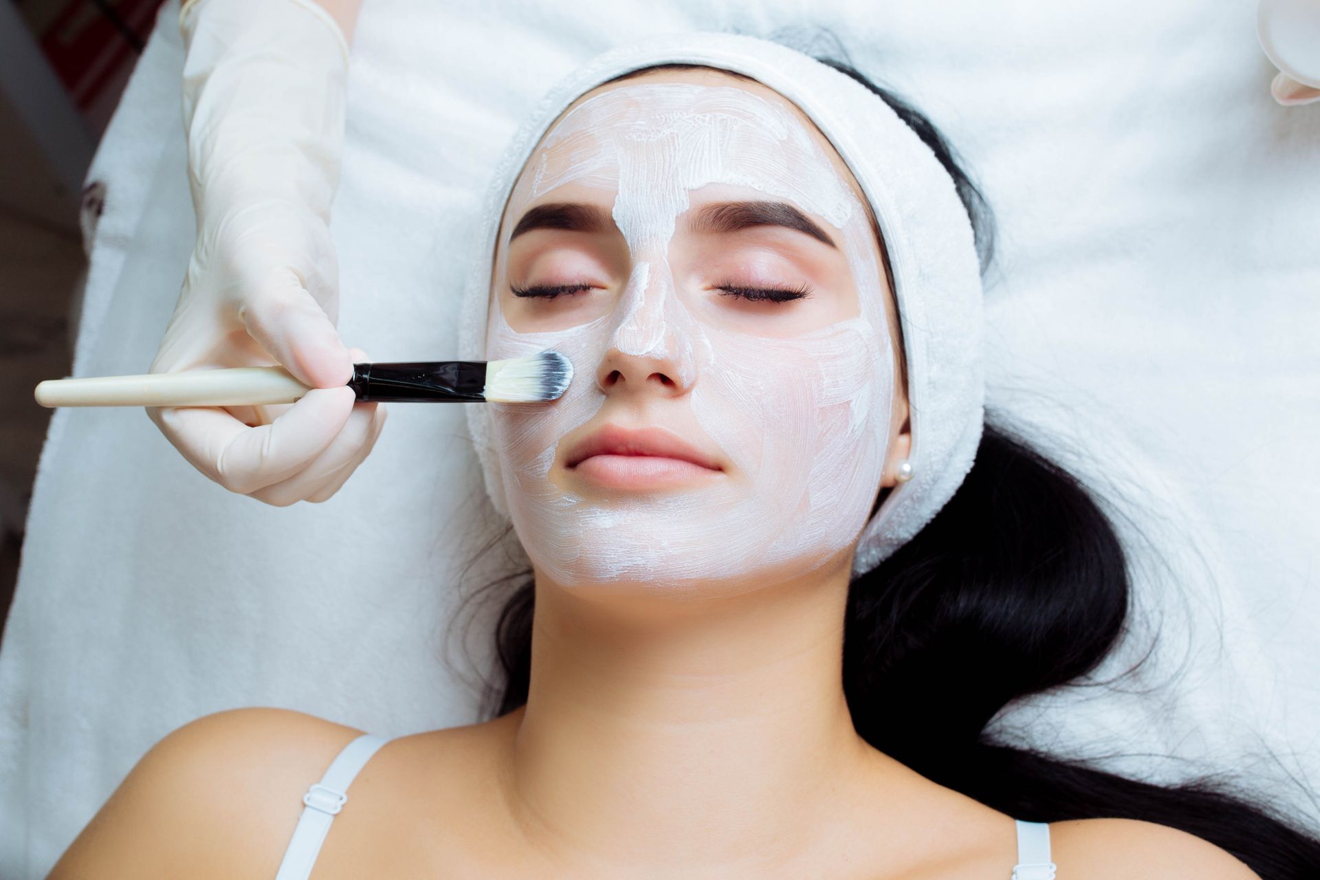 A gloved hand uses a brush to apply a white facial mask to a person lying on a spa bed with their eyes closed.