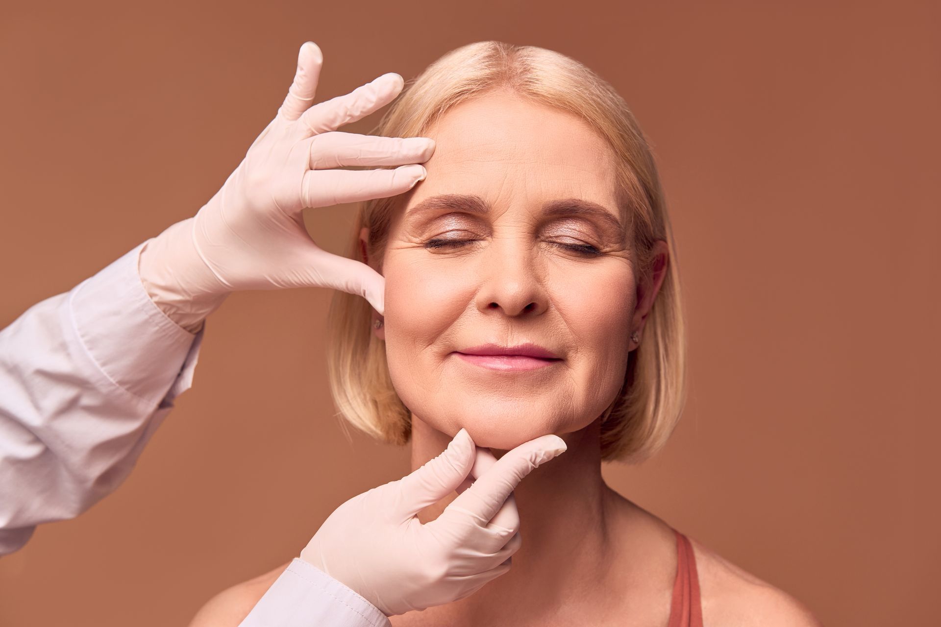 Cosmetic professional examining an older woman's face with gloved hands.
