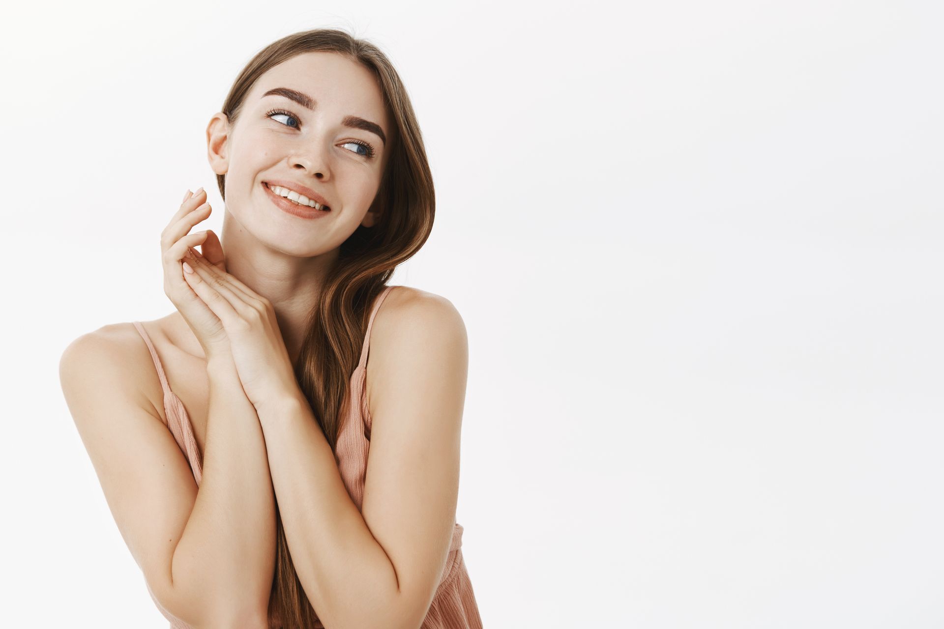 Woman with long brown hair, hands clasped near her face, smiling and looking up, against a white background.