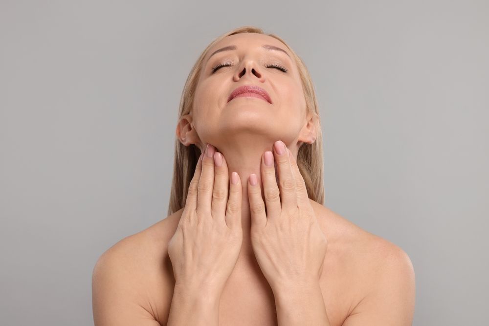 Woman with eyes closed, hands on neck, against a gray background.