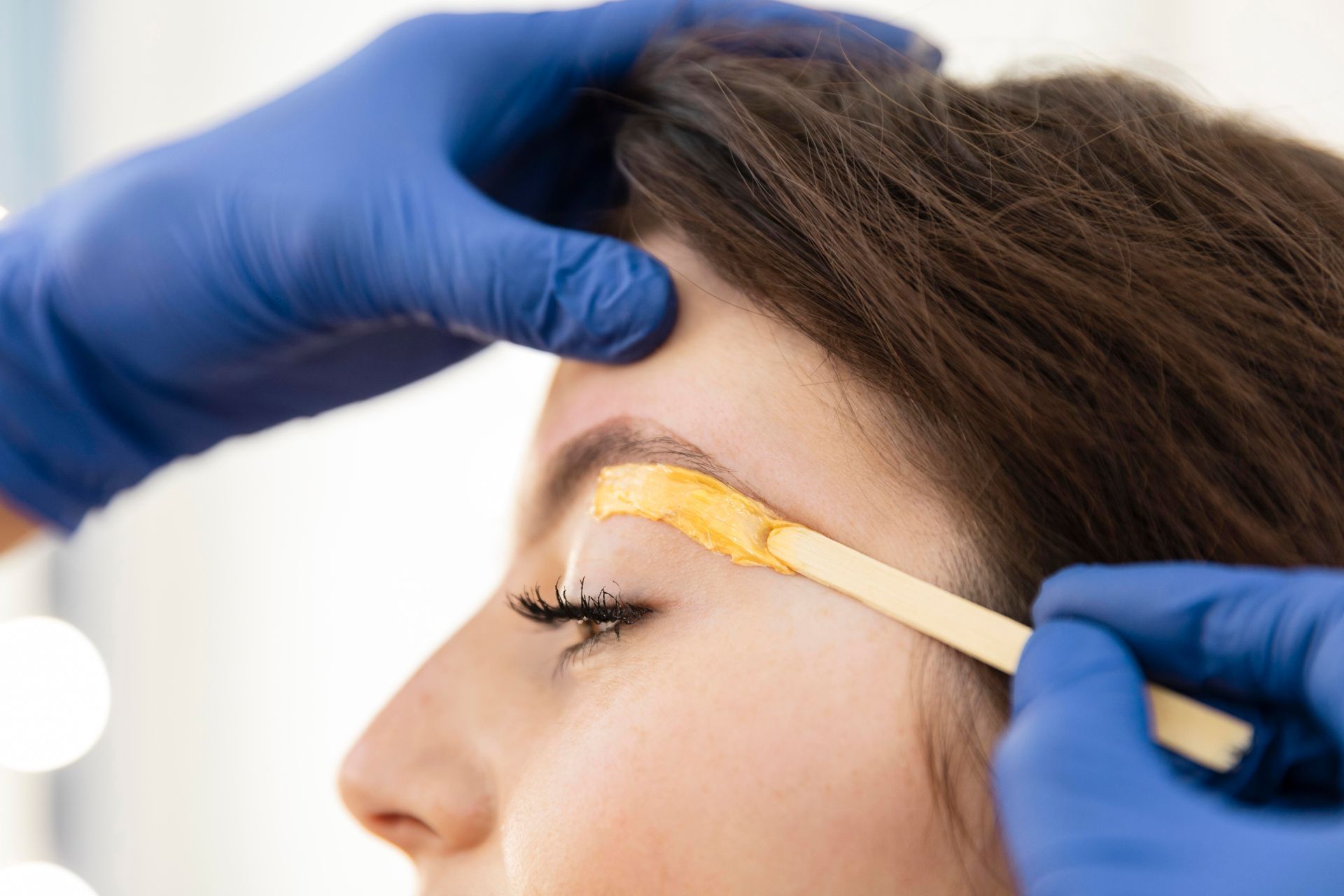 A woman is getting her eyebrows waxed by a person wearing blue gloves.