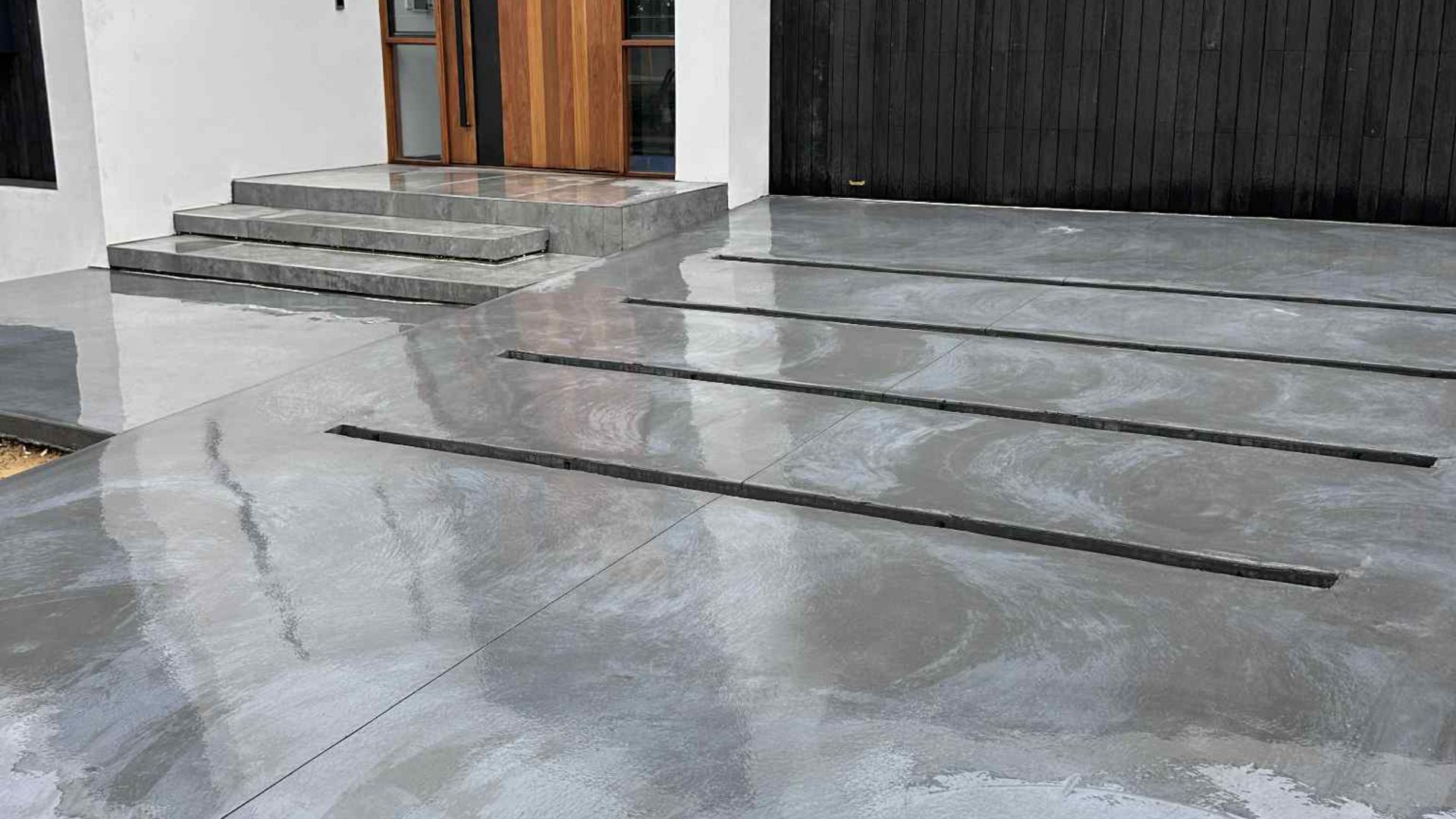 Grey concrete driveway with linear drain slots leading to a wooden door and steps.