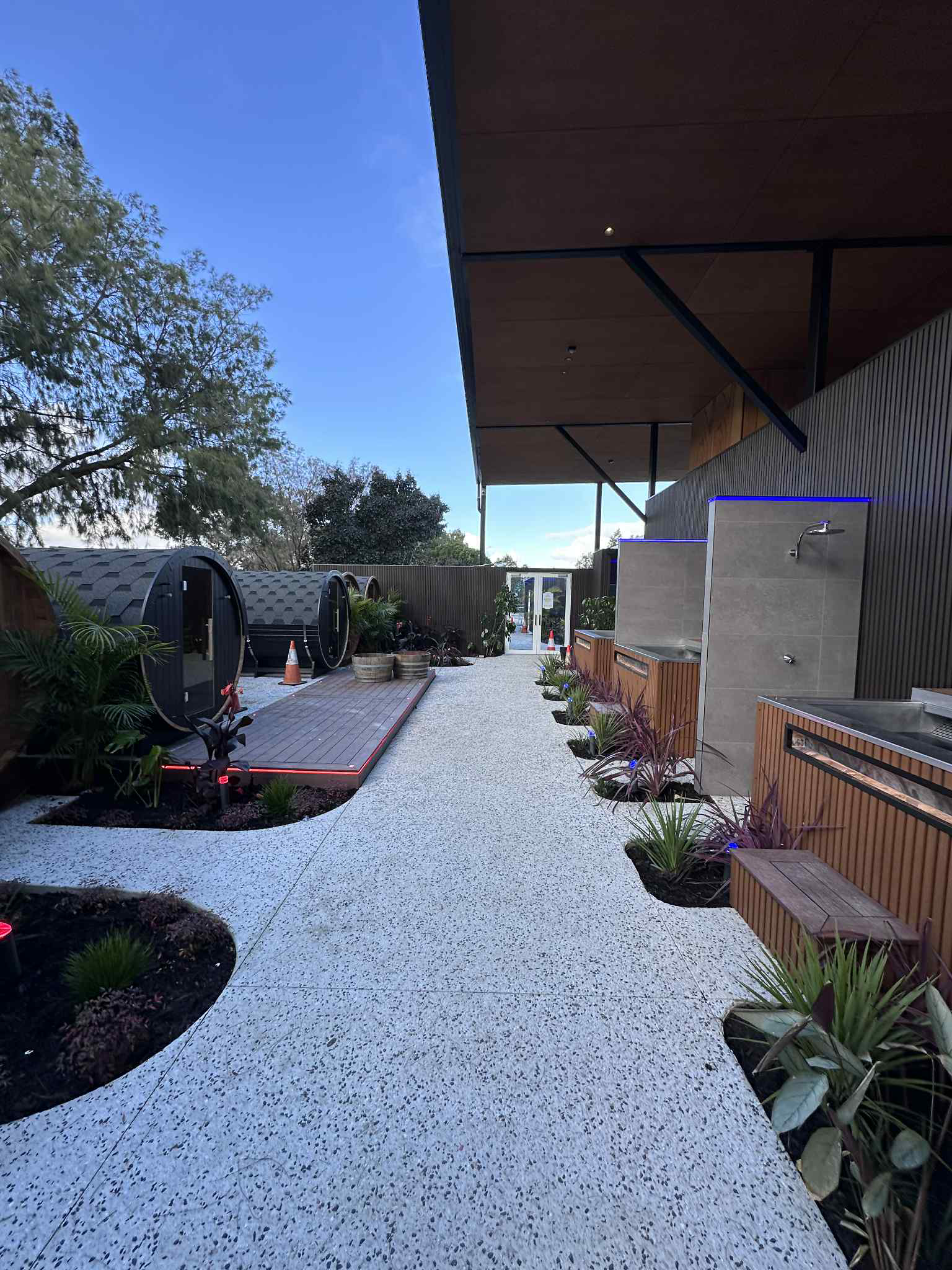 Pathway of white stones lined with garden features and outdoor showers. Wooden structures and barrel saunas visible.
