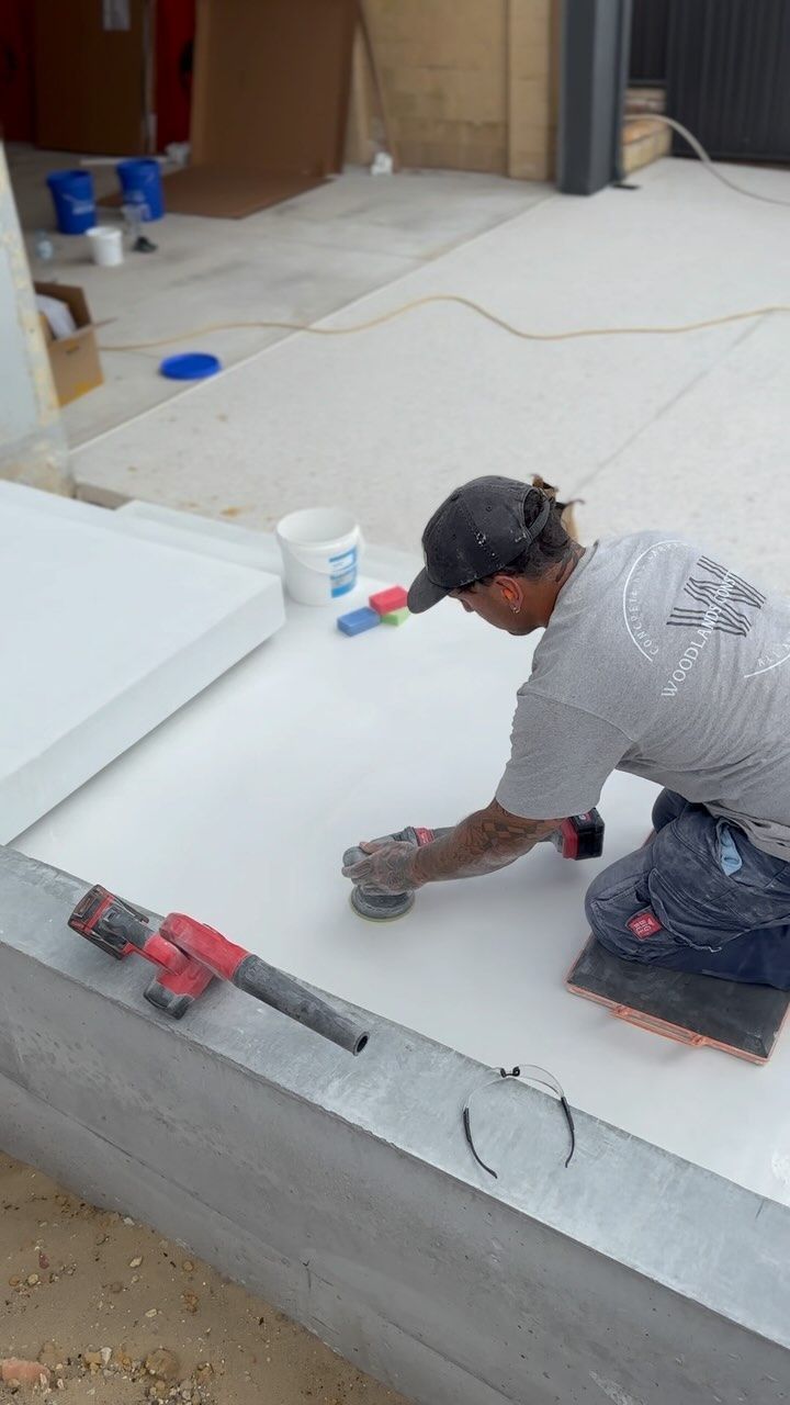  A man sanding a white concrete surface outdoors. He kneels, wearing a hat, using a power sander.