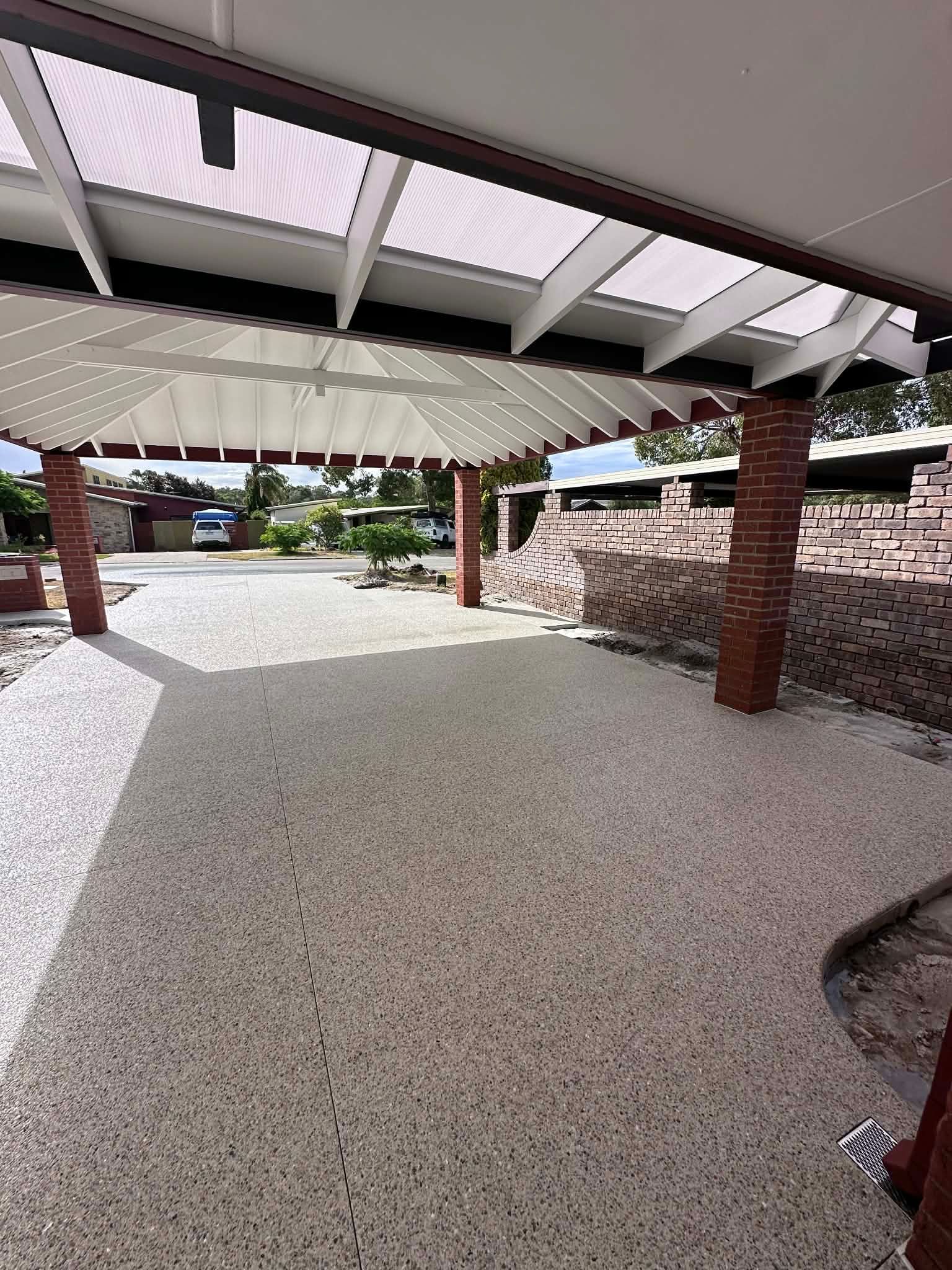 A covered outdoor area with a pebble floor, supported by brick pillars, white beams, and a slatted roof.