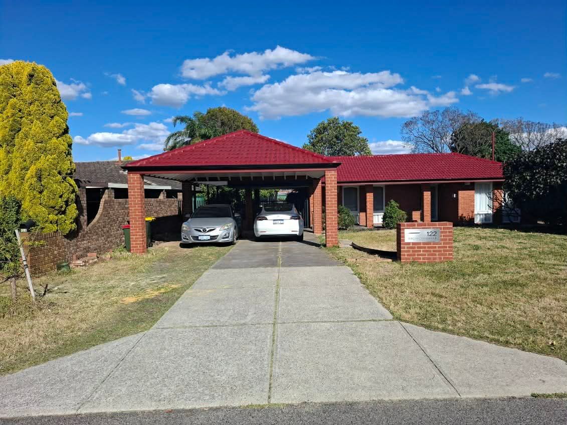Brick house with red roof, carport with two parked cars, and concrete driveway on a sunny day.