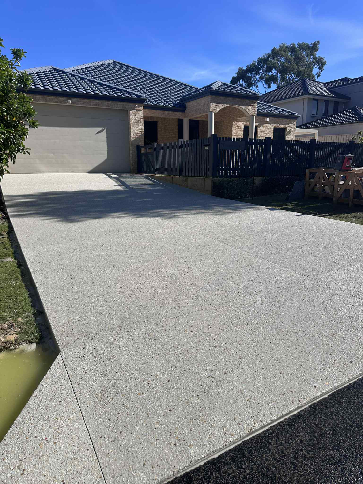 A gray aggregate driveway leads to a light brick house with a tiled roof, under a clear sky.
