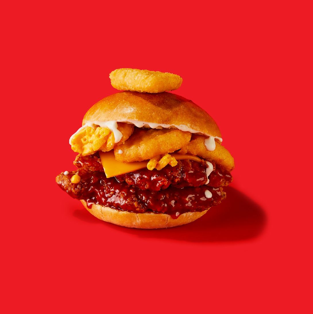 A chicken sandwich with cheese and onion rings on a bun on a red background.