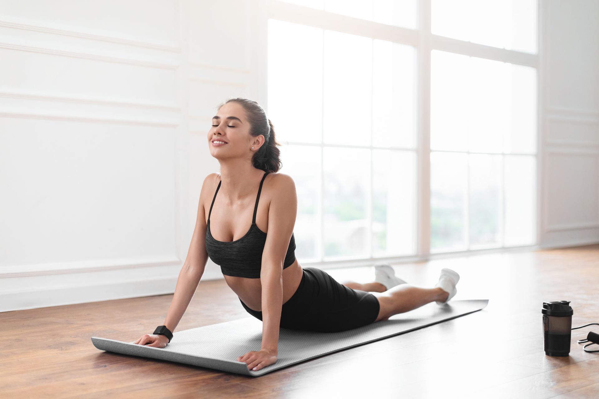 Woman in athletic wear doing yoga pose on a mat; bright room with large window.