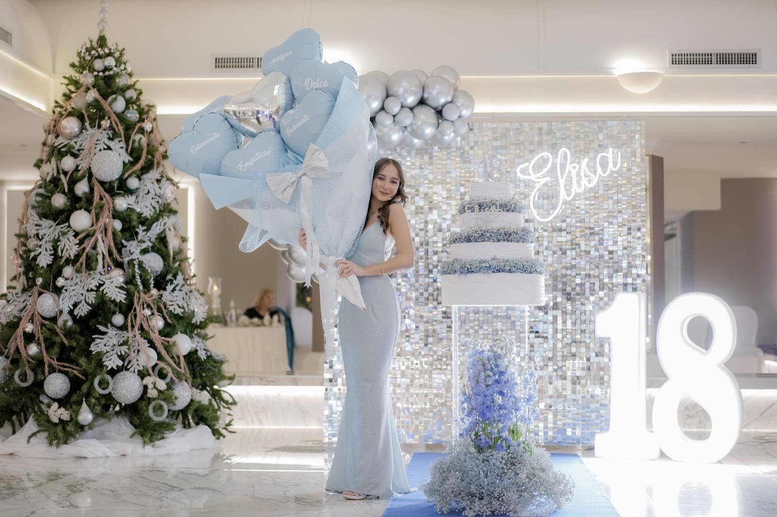 Una donna vestita di blu tiene in mano un mazzo di palloncini davanti all'albero di Natale.