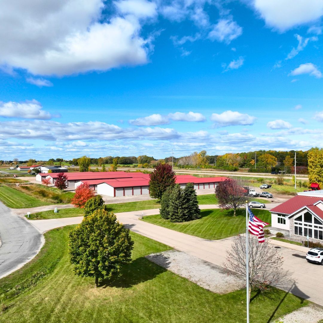 Oshkosh self-storage with red roofs against a blue sky, surrounded by green grass and trees. American flag in the foreground.