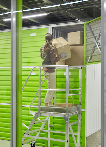 A woman is standing on a ladder in a warehouse holding a box.