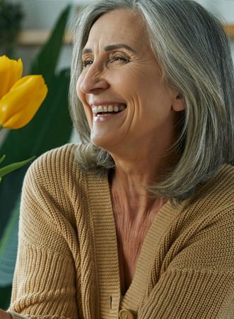 A woman with gray hair is smiling while holding a yellow flower.