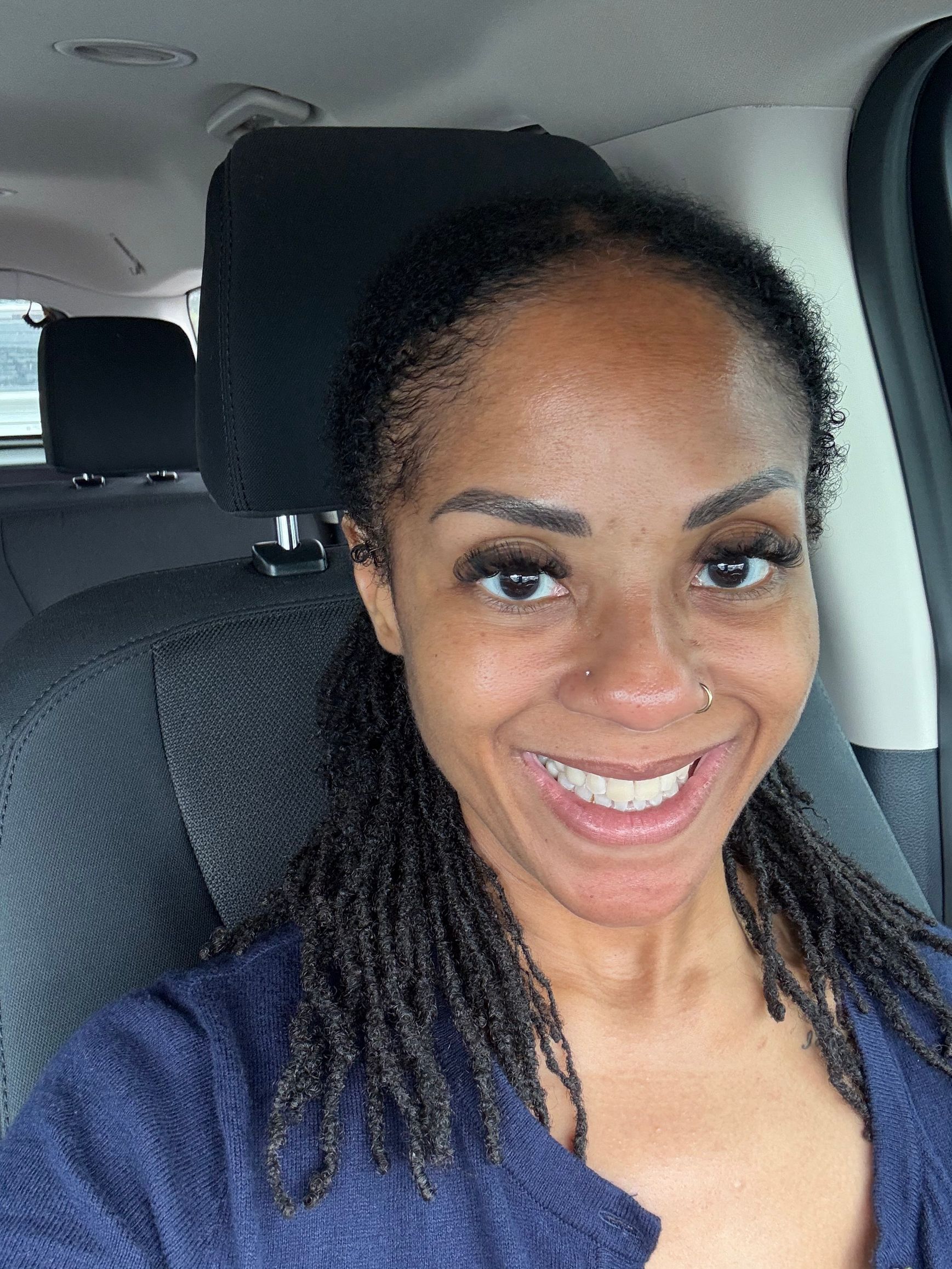A smiling Black woman with locs sits in a car, wearing a blue sweater.