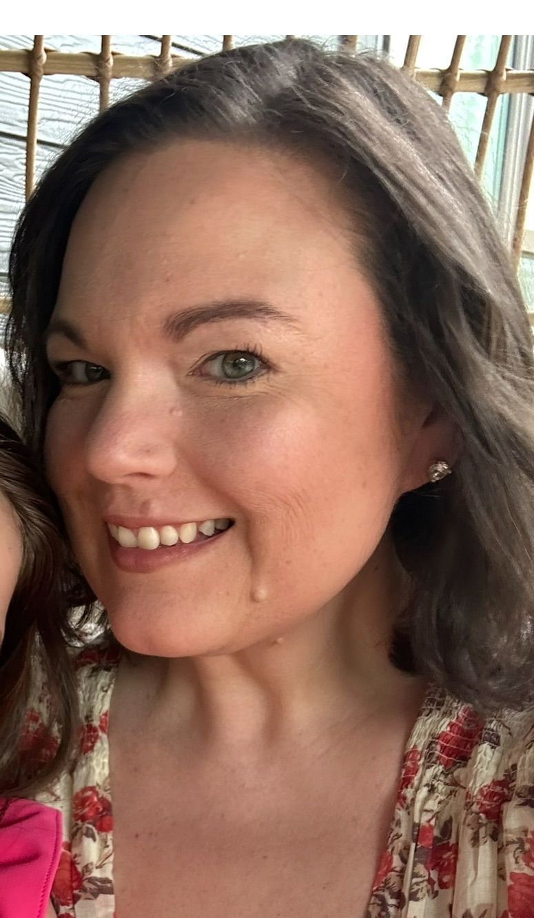 Woman with brown hair and a pink floral top smiles; she has a teardrop on her cheek.