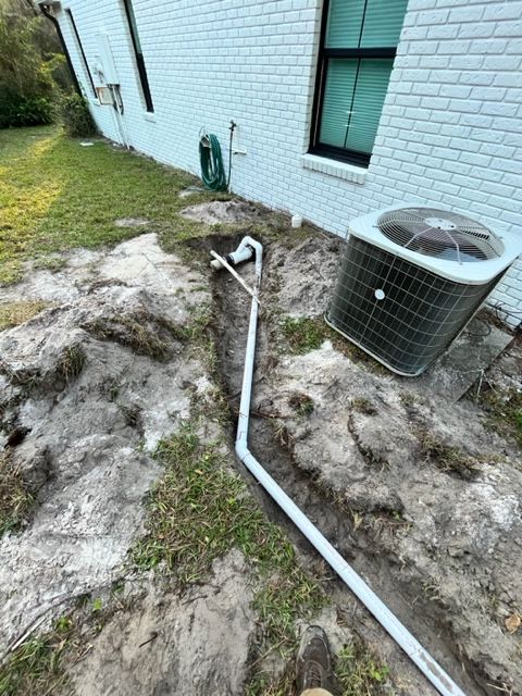 A grey pipe runs through a dug trench in the ground alongside a white brick house near an outdoor AC unit.