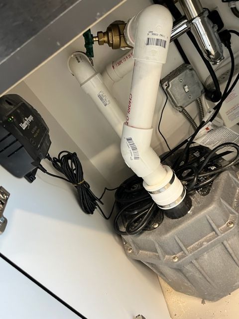 A gray sewage ejector pump system with white PVC discharge piping and a black power alarm box inside a cabinet.