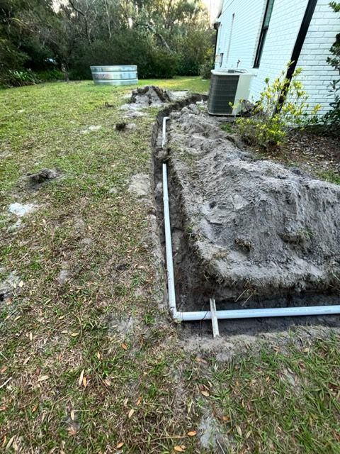 A trench with white PVC drainage pipes being installed along the side of a white house in a grassy yard.