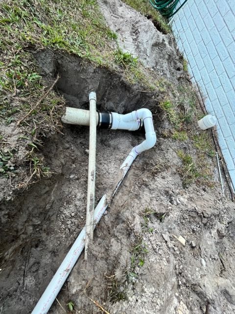 An outdoor trench shows a broken white PVC drain pipe connected to a new white PVC elbow with a black rubber coupling.