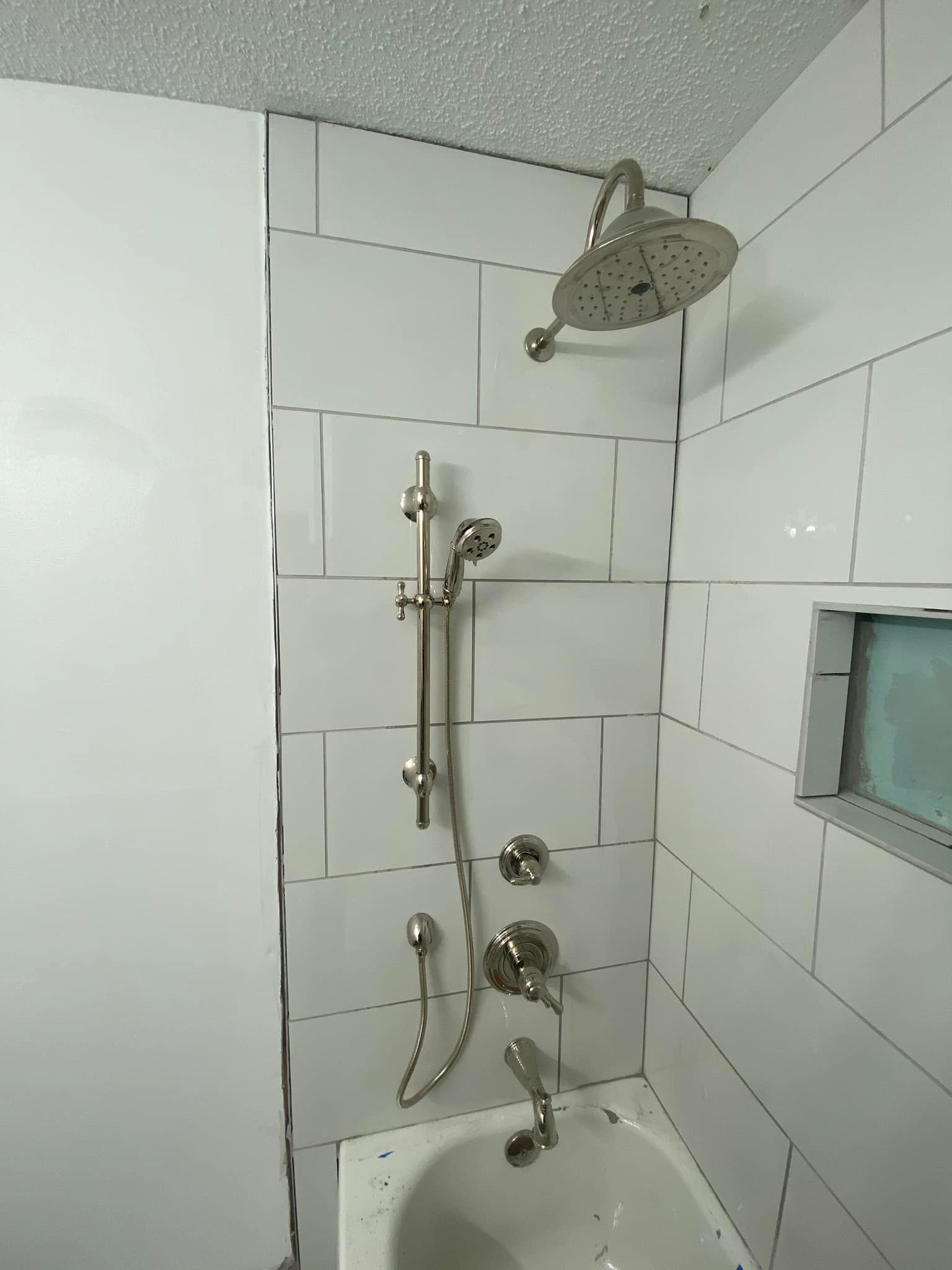 A white tiled shower featuring a ceiling-mounted rainfall showerhead, a slide bar hand shower, and a bathtub faucet.