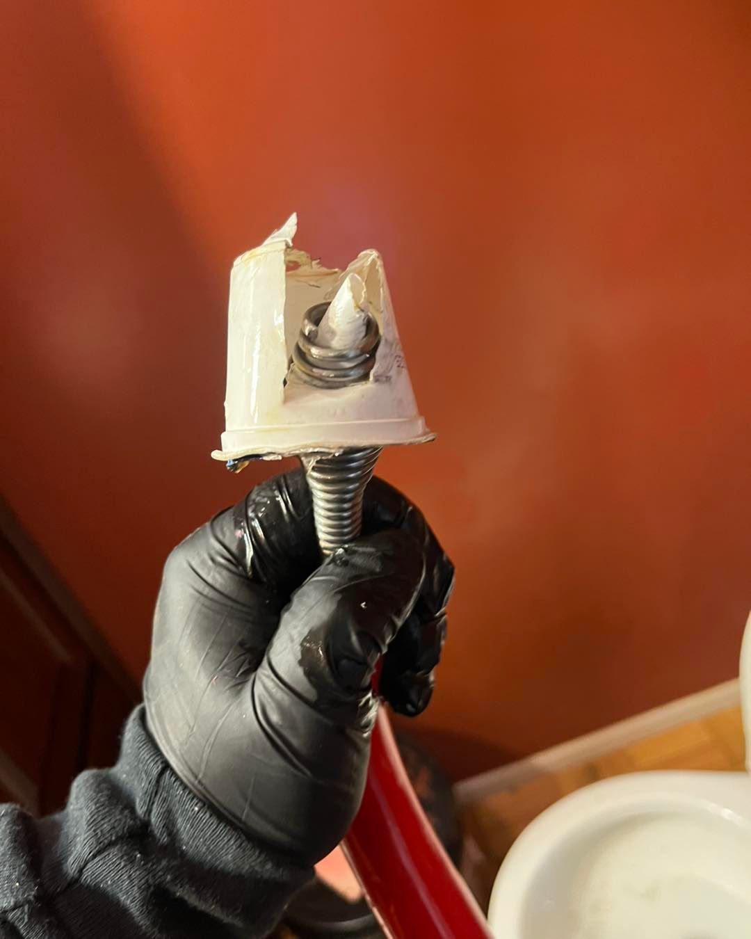 A gloved hand holding a damaged plastic toilet supply line connector with a broken plastic nut and exposed metal threads.
