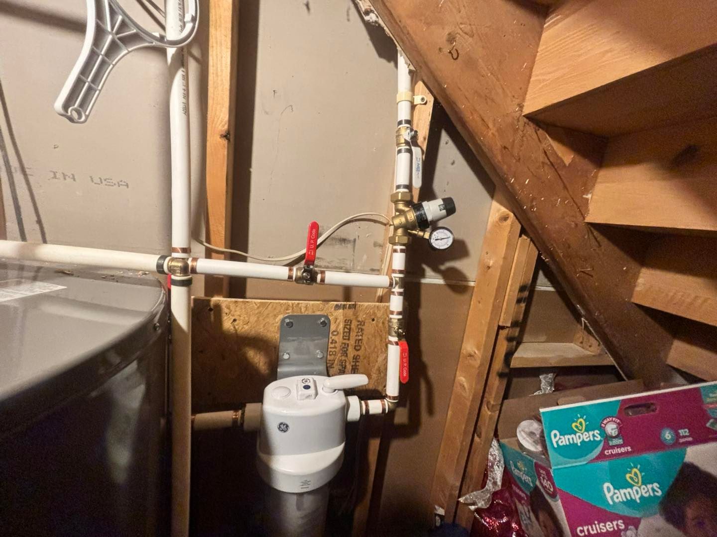 Plumbing setup with a white water filter housing, brass pressure regulator with gauge, and shut-off valves near stairs.
