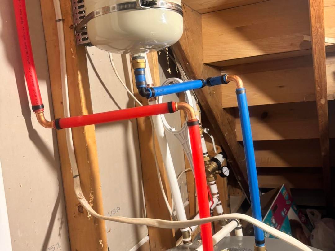 A metal expansion tank connected by blue and red PEX plumbing pipes installed against wooden wall studs.