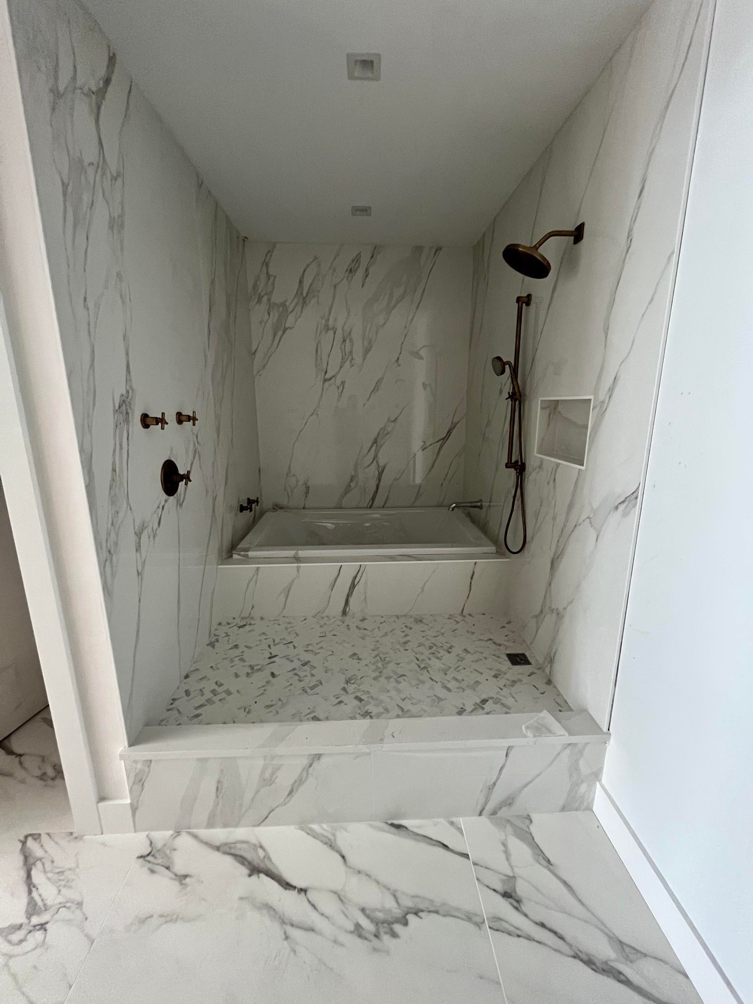 A walk-in shower area features white marble-look tiles with grey veining, a built-in tub, and bronze fixtures.