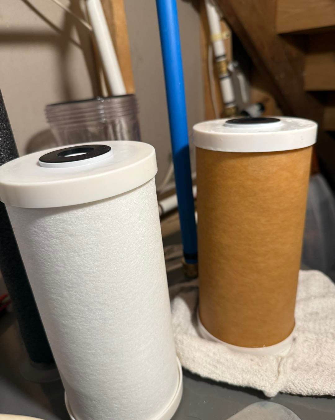 A new white water filter cartridge sits next to a used, discolored brown one on a surface near plumbing pipes.