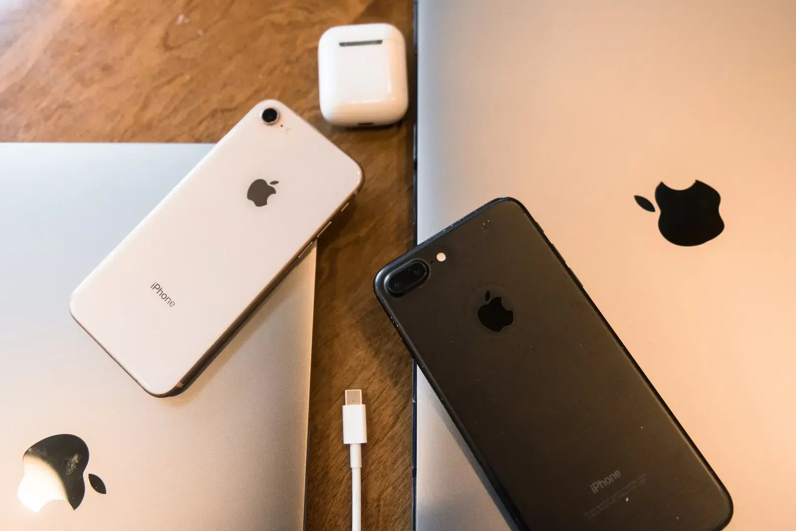 Three Apple devices on a table: white iPhone, black iPhone, AirPods case, and a MacBook with Apple logos
