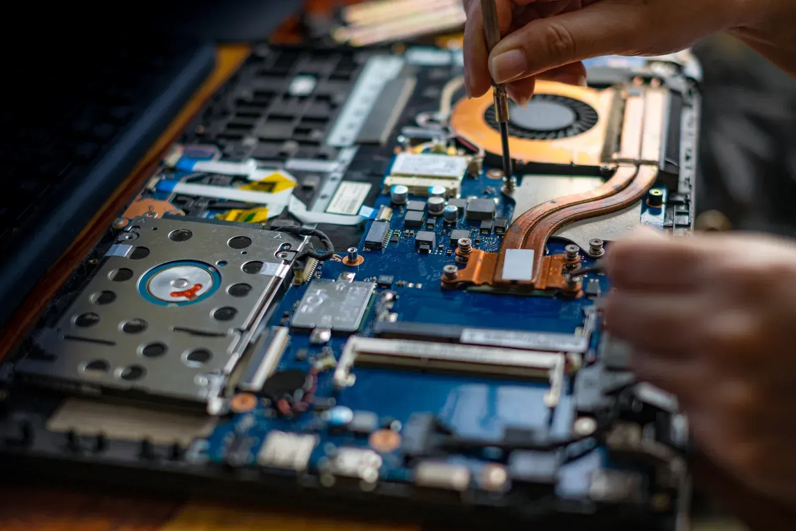 Hands repairing a laptop motherboard with a screwdriver and copper cooling strip