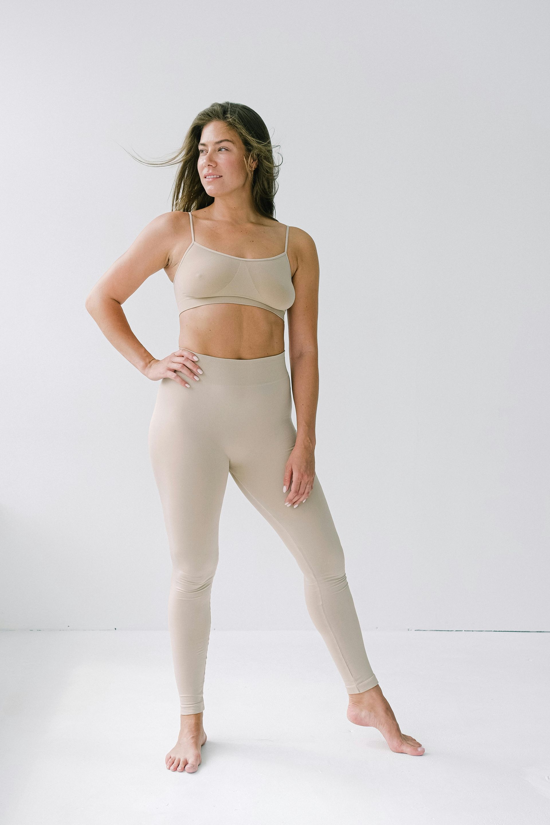 Una mujer con sujetador y leggings beige posa sobre un fondo blanco.