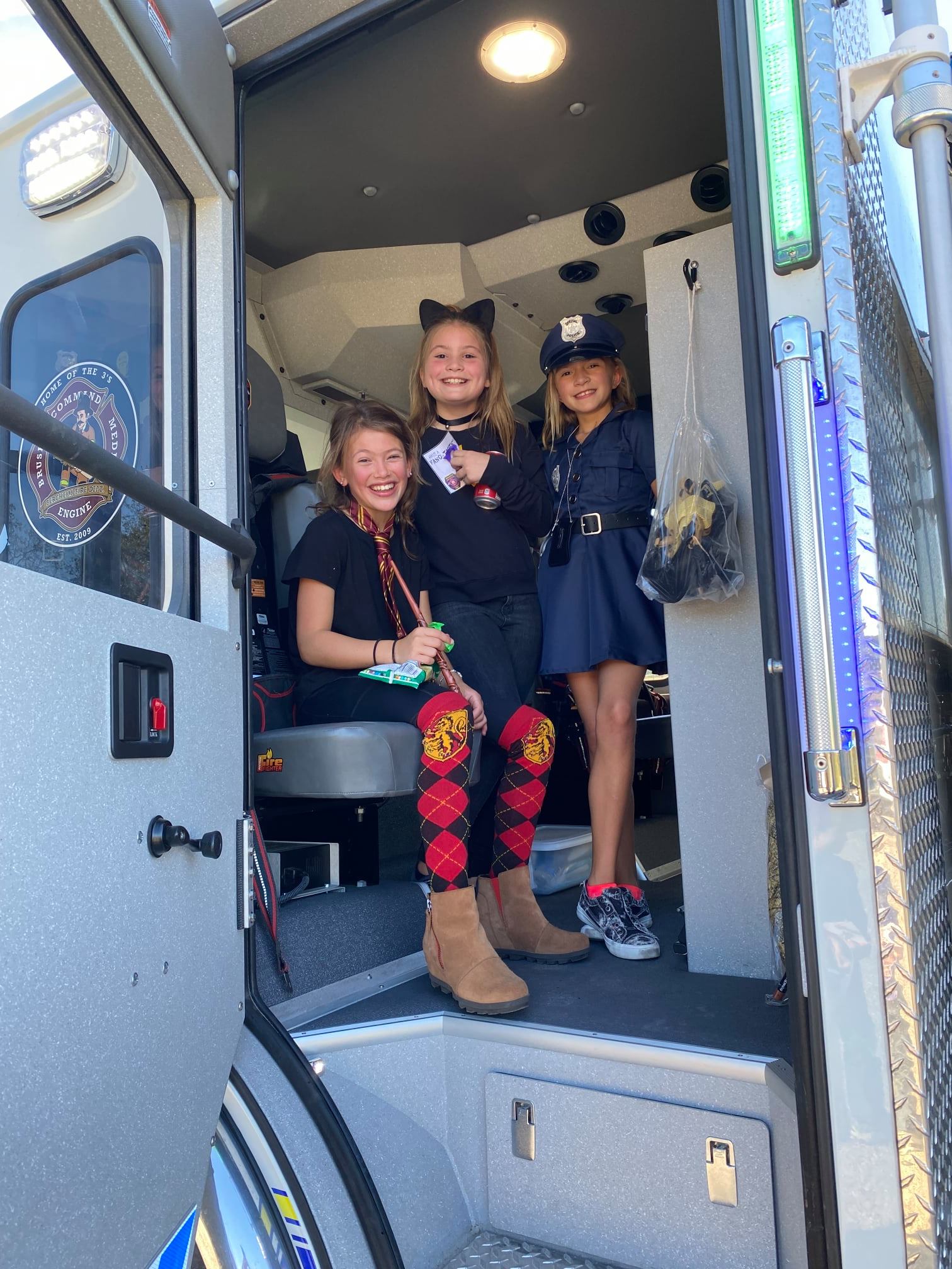 Three young girls in costumes are standing in the doorway of an ambulance.