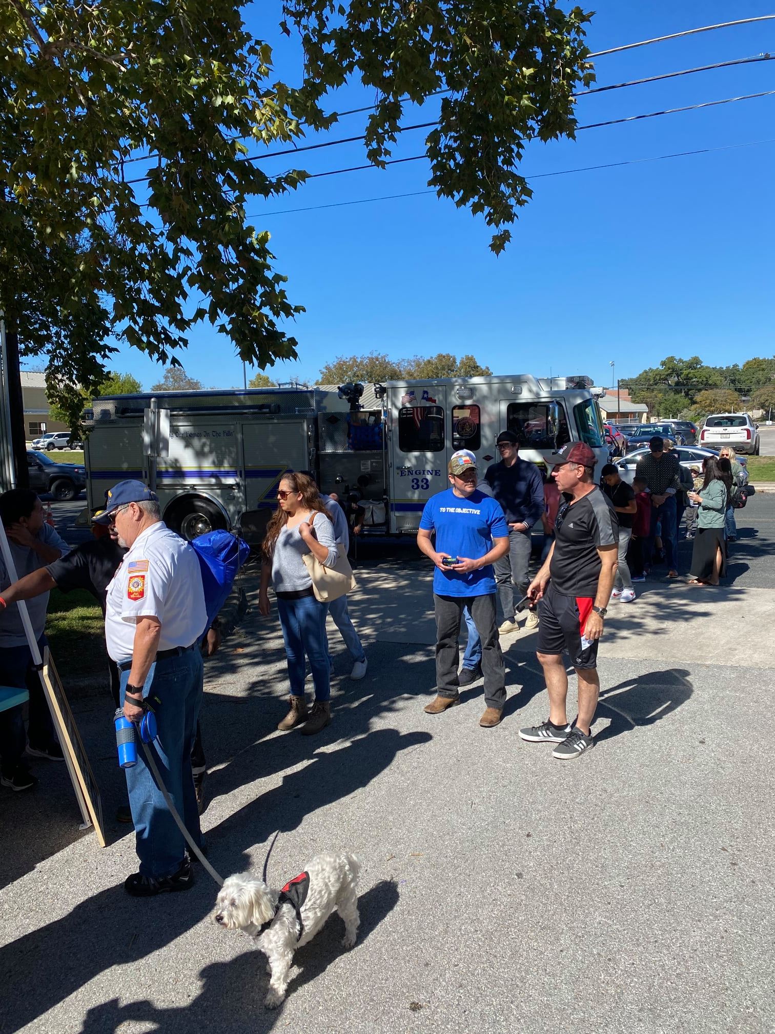 A group of people standing in a line with a dog on a leash in front of a whiter fire truck.