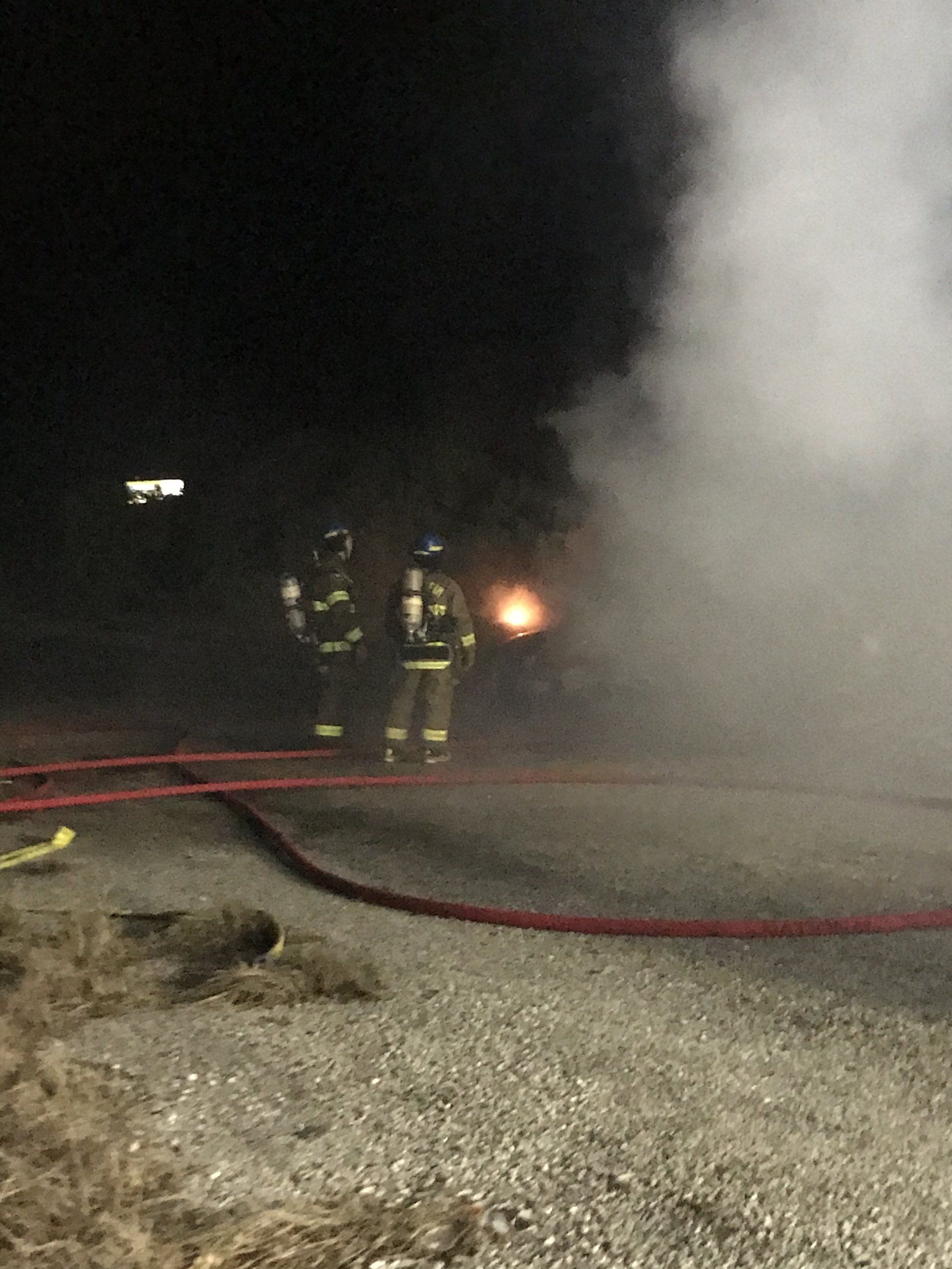 A pair of fire fighters are watching a training fire burn with fire hoses on the ground.